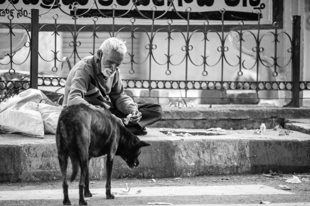 Black and white image. Older-appearing man in in background, shovelled clothing and hygiene. Man is sitting on cement curb with rot-iron fence at his back. Dark colour dog in fore-ground with head down, cautiously approaching man. Dog is facing aware from image viewer, with tail down neck lowered and ears perked at sides of head.
