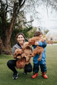 Mother kneeling on grass beside young child. standing, cuddling curly haired, brown dog. Mother is holding 2 more curly haired brown dogs. Dog companions enhance the lives of autistic children. 