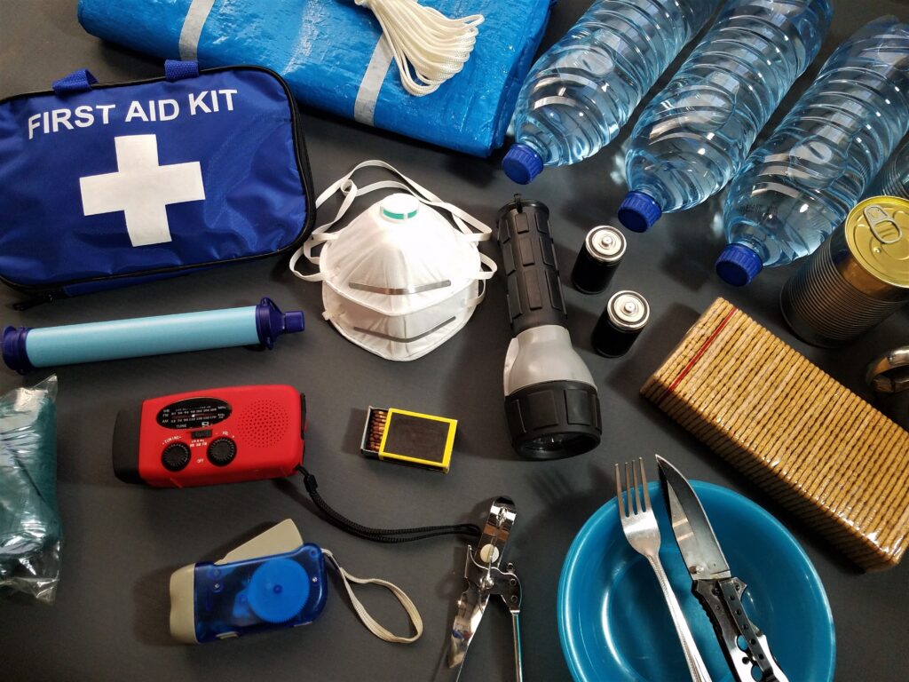 multiple tools and dressings of a first aid kit are scattered about. Including flashlights, batteries and bottles of water. A noticeable blue pack is in the upper left corner of image, reads "first aid kit".