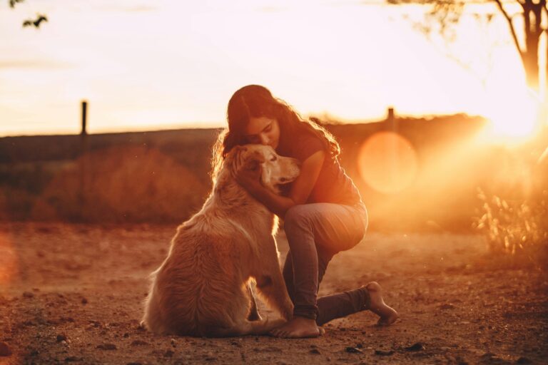 Lady kneeling down to embrace large beige dog, with sunset in the background.